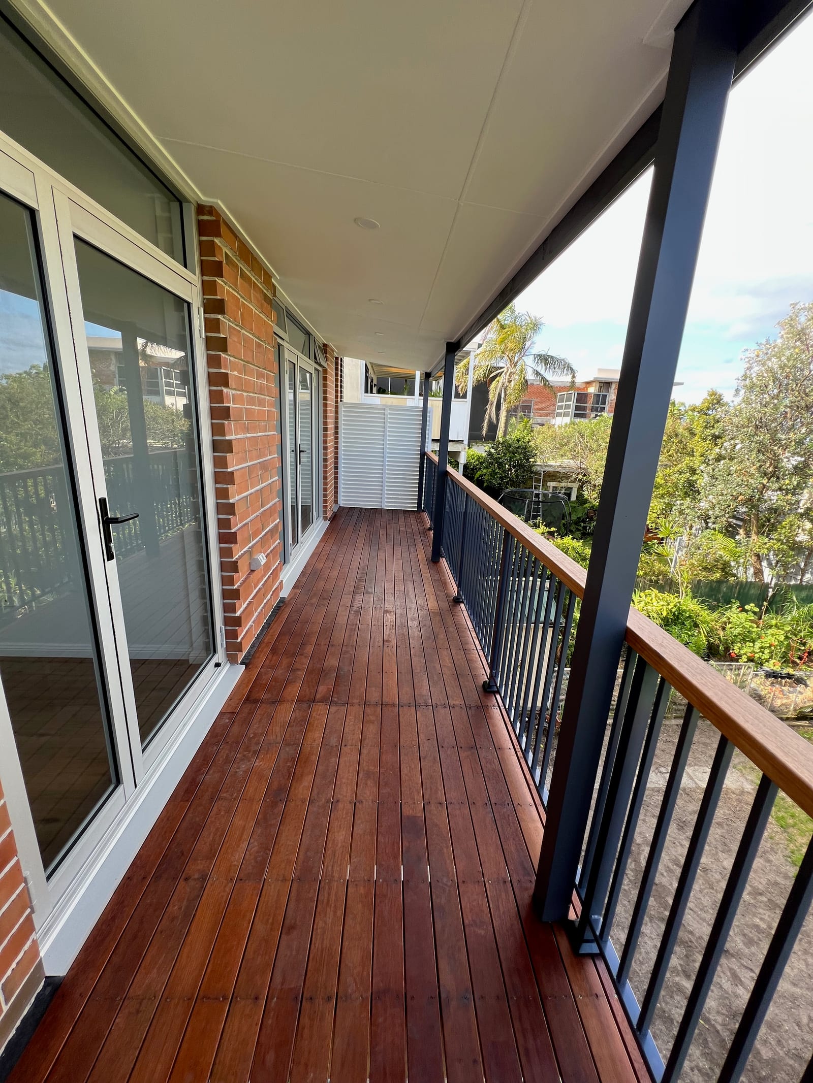The finished upper-level balcony — timber decking, a steel balustrade with timber handrail, sliding glass doors, a brick pier and a louvred privacy screen