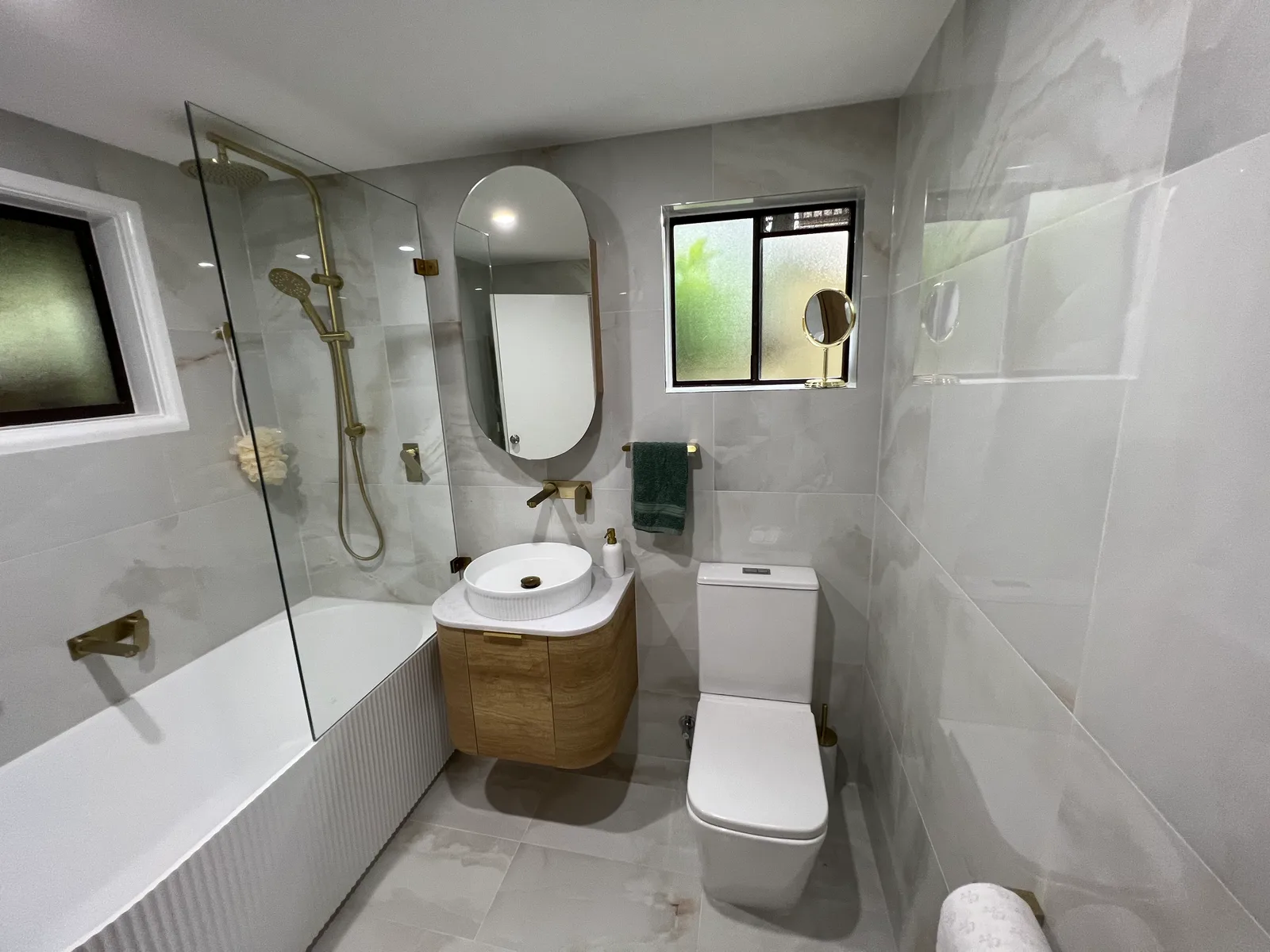 A bathroom with a freestanding-style bath behind a frameless glass screen, an oval mirror, brushed-brass tapware and a round stone basin on a timber vanity.